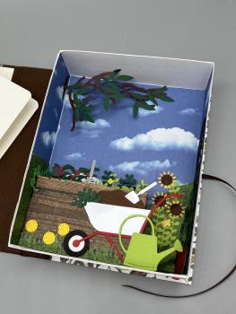 Preview: BuchBox Gartenarbeiten Variante Hochbeet – geöffnet, Garten-Szenerie mit Schubkarre und Gießkanne