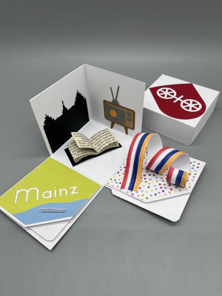 Explosionsbox Mainz geöffnet – Szenerie mit Fastnachtsband, Gutenberg-Minibuch und altem Fernseher aus Papier